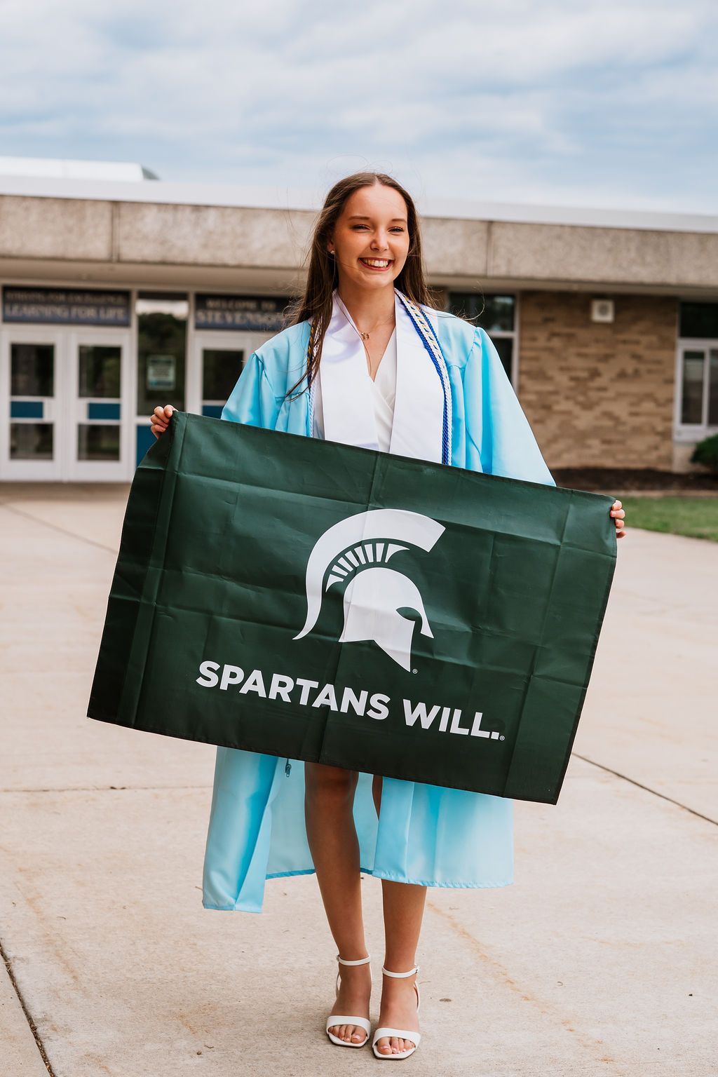 Graduate in light blue graduation gown holds Michigan State Spartans flag outside campus building.