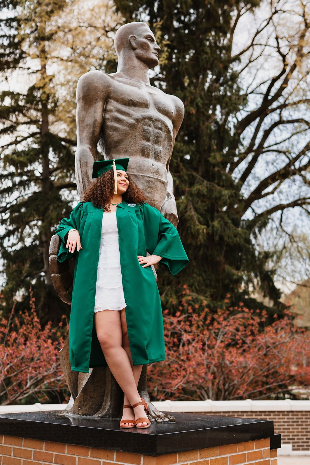 A person in a green blazer and white dress sits casually on a brick wall near flowering trees in spring.