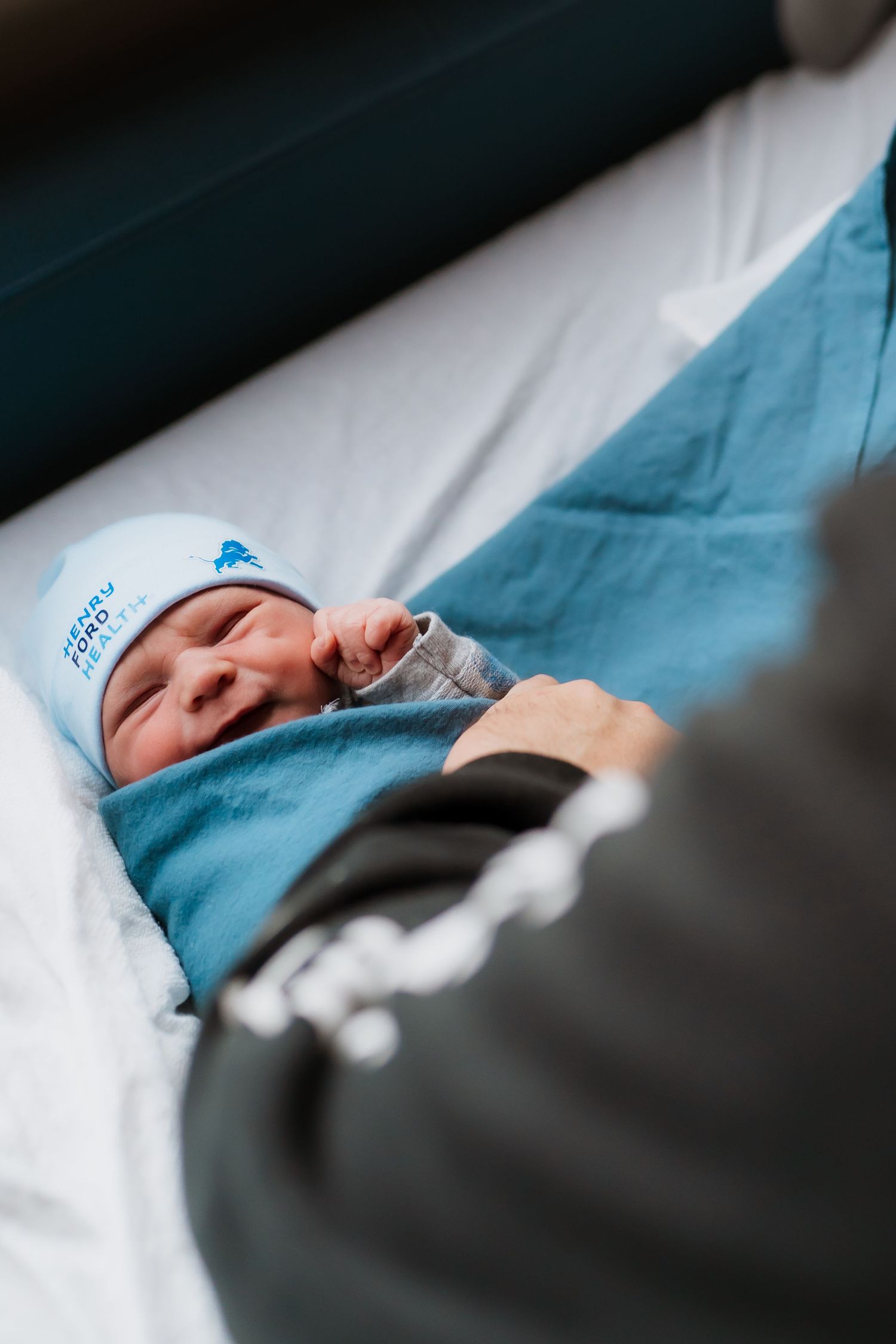 Newborn baby wrapped in blue blanket wearing a Detroit Lions hat lying on a white hospital bed.