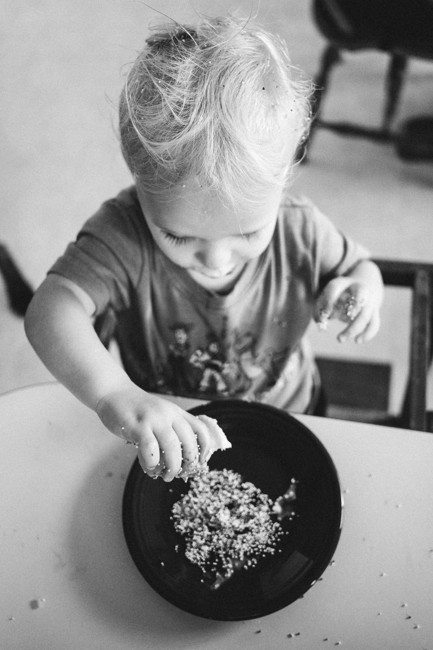 Smiling toddler playing with rice in a black bowl at a table, black and white photo.