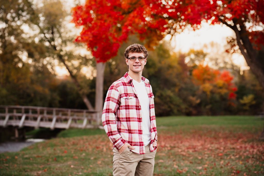 Young man in plaid flannel shirt stands in autumn park with vibrant red foliage and wooden bridge backdrop.