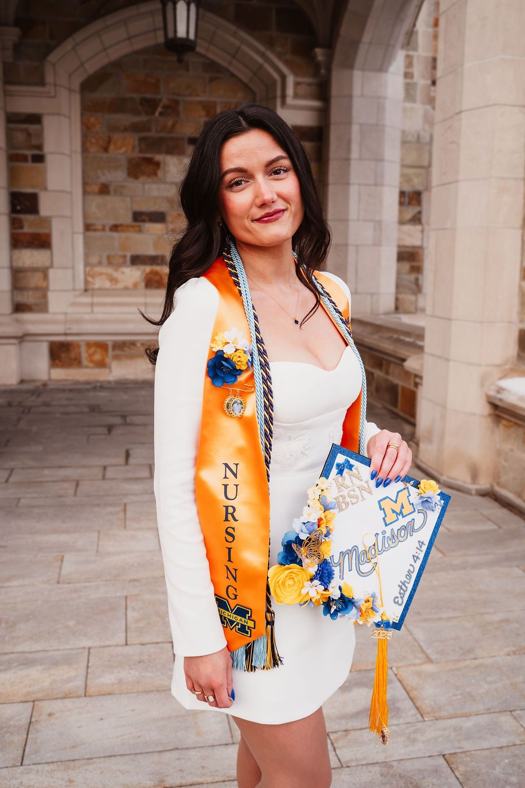 College graduate wearing white top and orange graduation stole holds diploma and decorative cap in stone archway.
