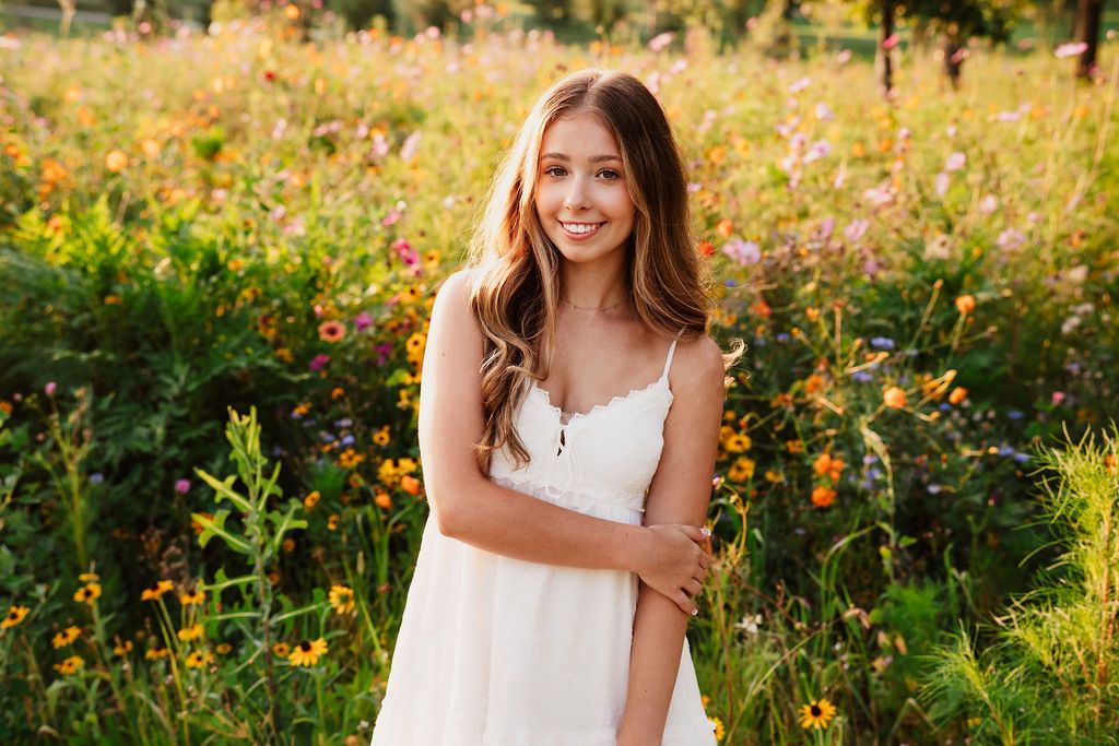A person in a white summer dress smiles while standing in a golden wildflower meadow during sunset.