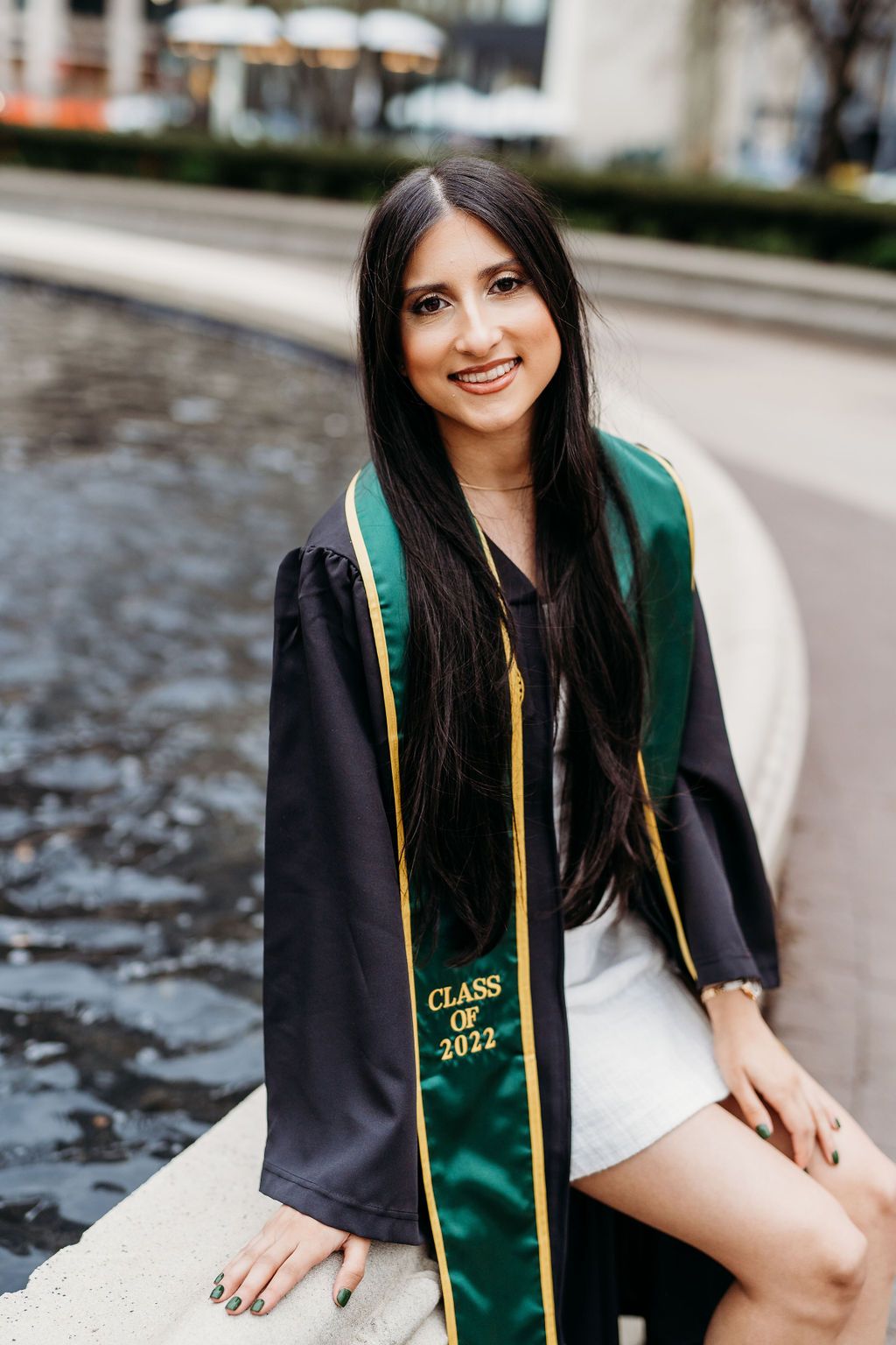 A graduate wearing a green and gold stole with black graduation gown sits outdoors for portraits.