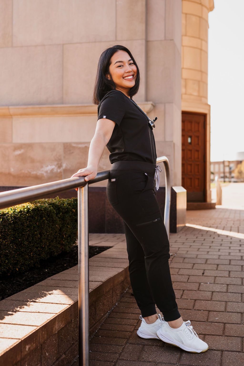 Graduate poses in a black graduation gown and white sneakers on brick pavement near stone columns.