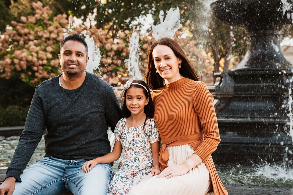A family of three smiles together by a fountain, featuring a man, young girl, and woman in warm autumn attire.