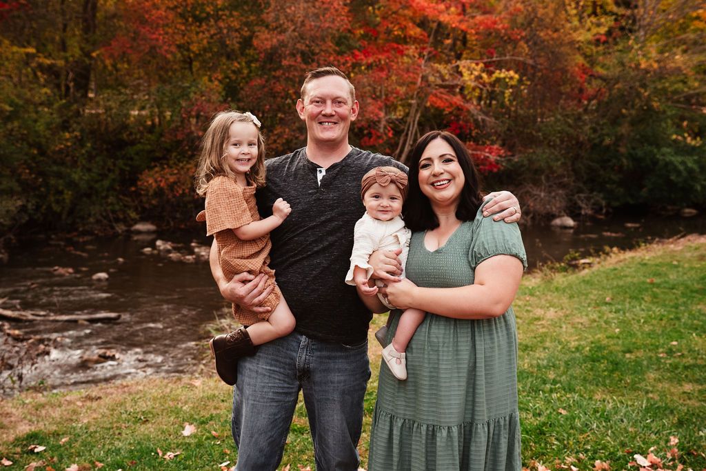Family of four posing outdoors in fall foliage near a creek, parents holding two young children, smiling warmly.