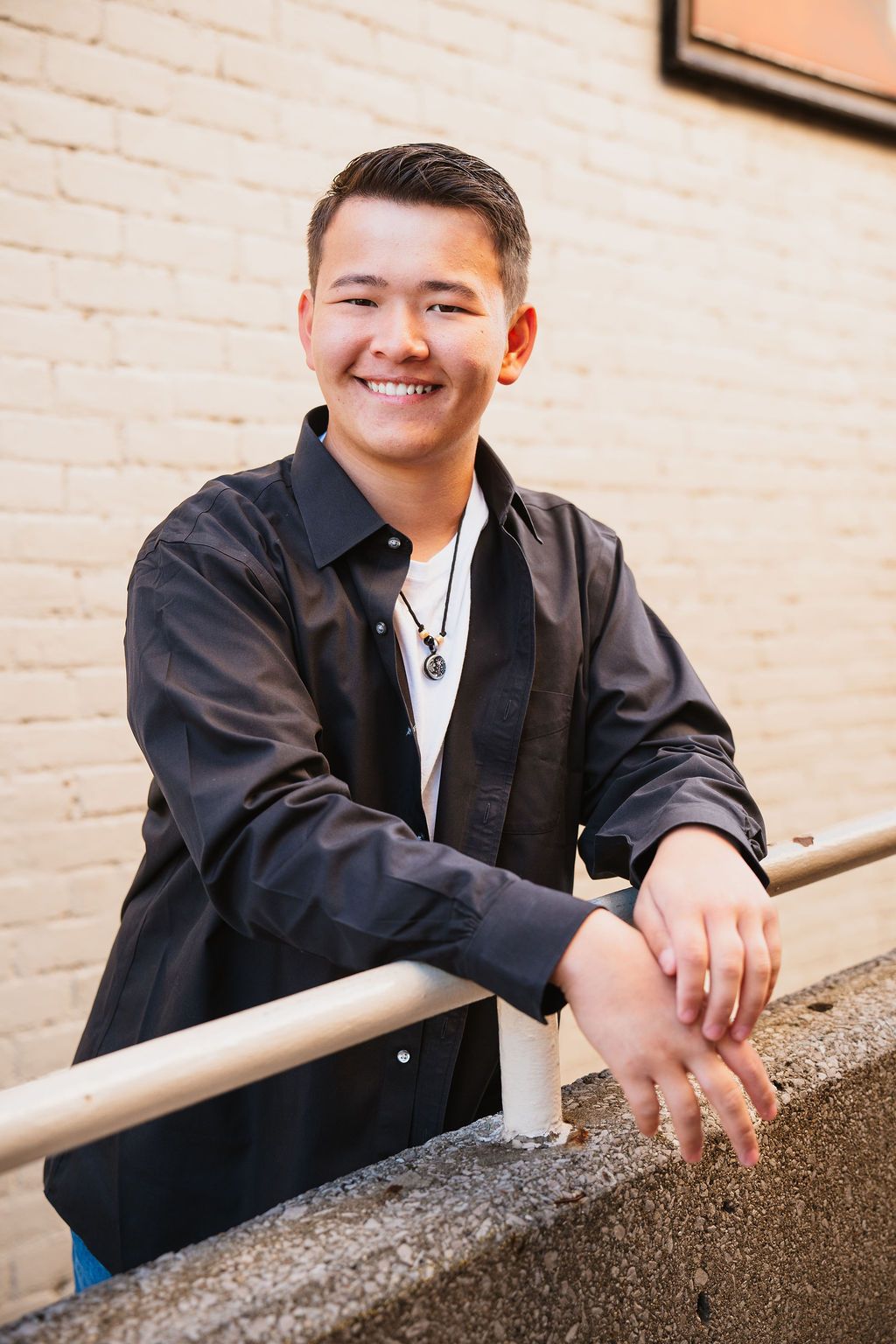 Smiling young Asian man in dark shirt leaning on metal railing against brick wall wearing necklace.