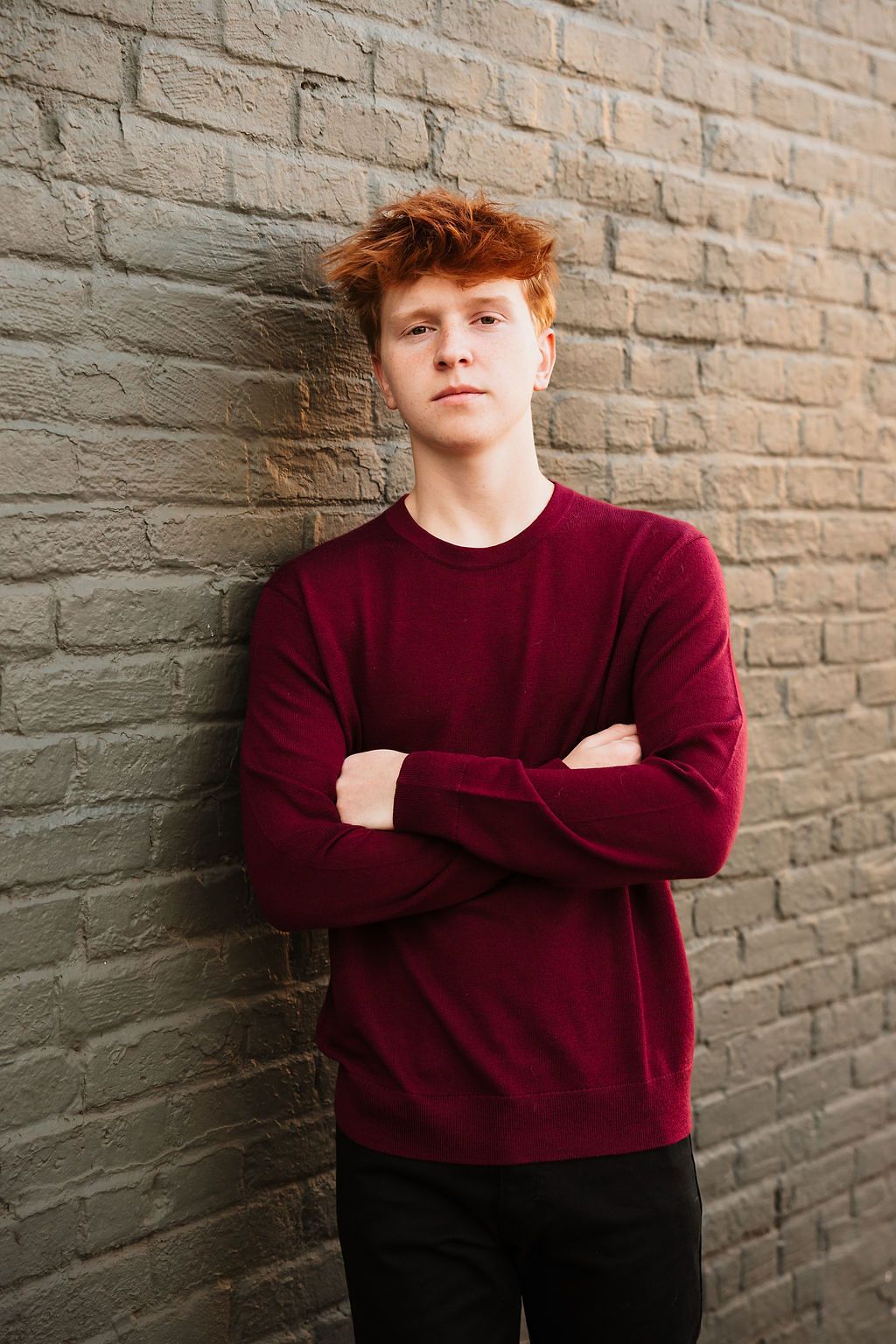 Young redhead man in burgundy sweater leaning against brick wall with arms crossed, confident pose.