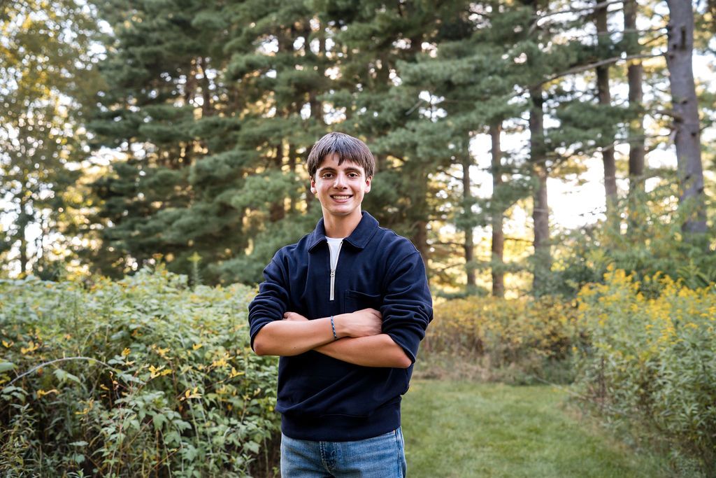 Smiling young man standing with arms crossed in a lush green forest park setting on a sunny day.