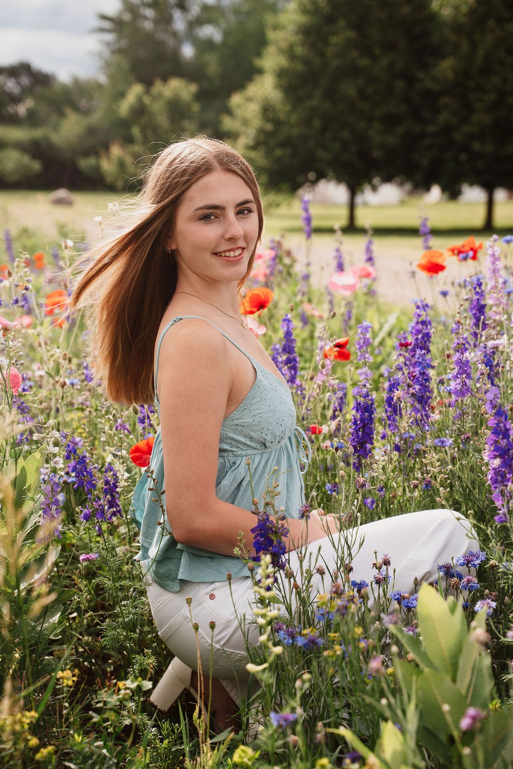 Young woman sitting in a vibrant wildflower field with purple and red blooms, wearing a teal top and white pants.