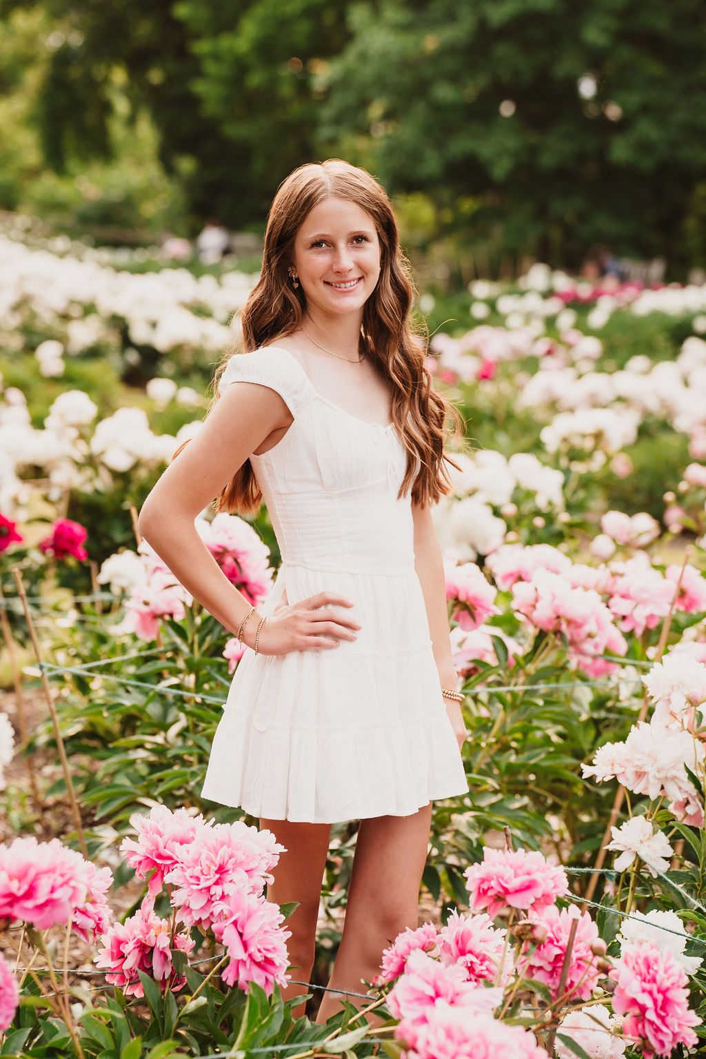 Young girl in white dress smiling among pink and white peonies in a lush garden.
