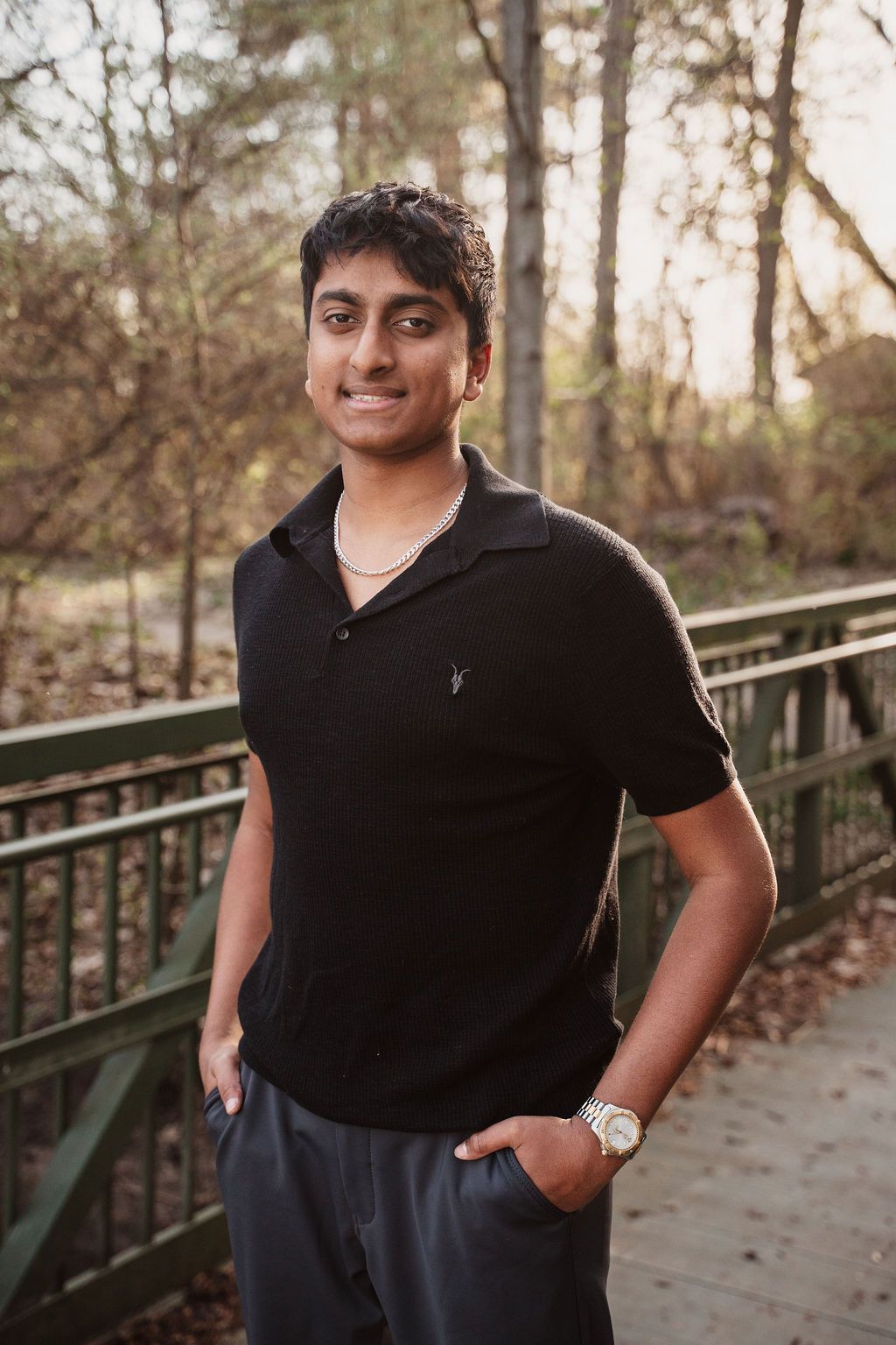 Young man in black polo shirt smiling on a wooden bridge surrounded by trees in a natural outdoor setting.
