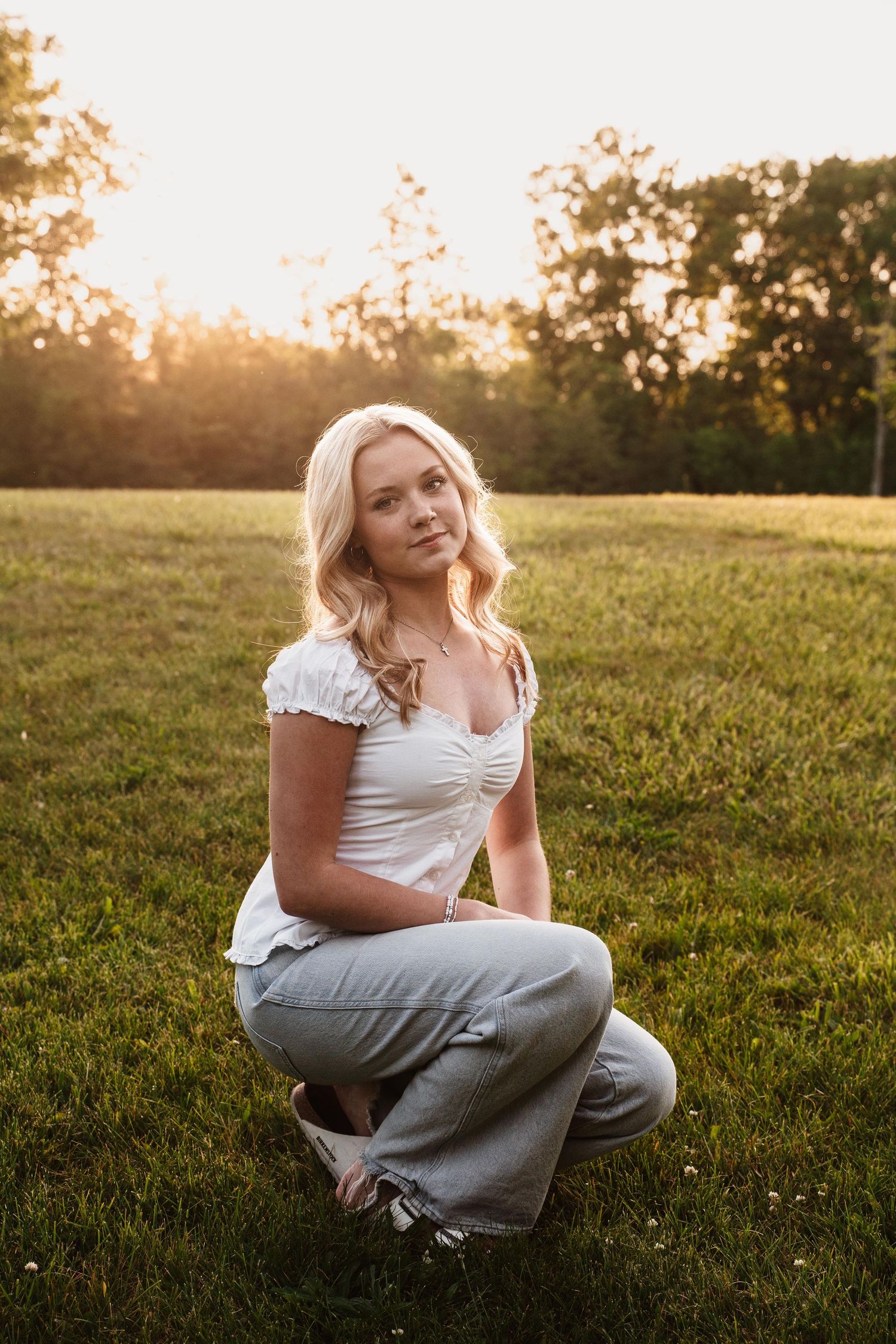 Blonde woman in white top and gray jeans sitting on green grass during golden hour sunset outdoors.
