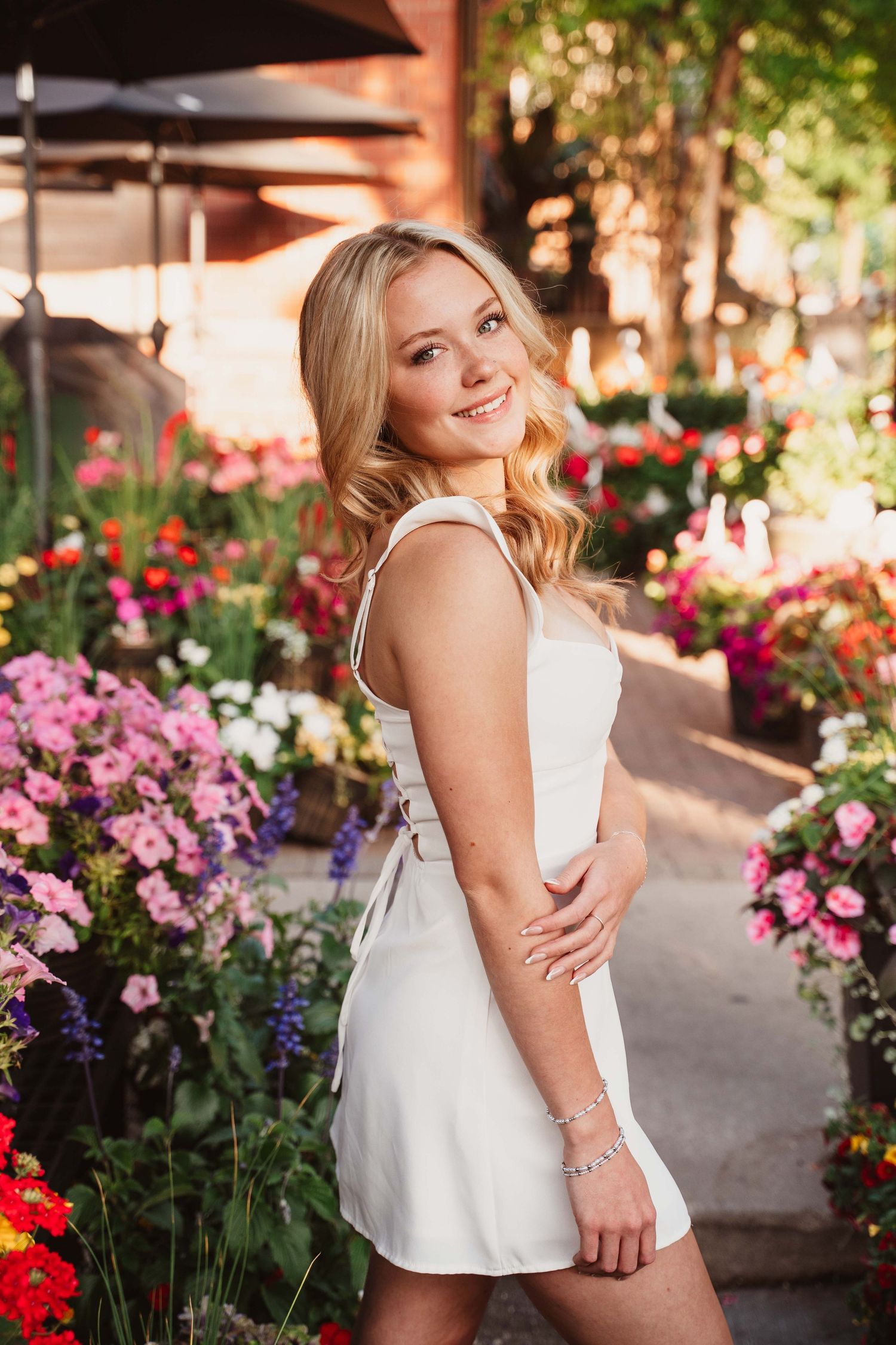 Smiling blonde woman in white dress posing among colorful flowers in a sunlit garden.