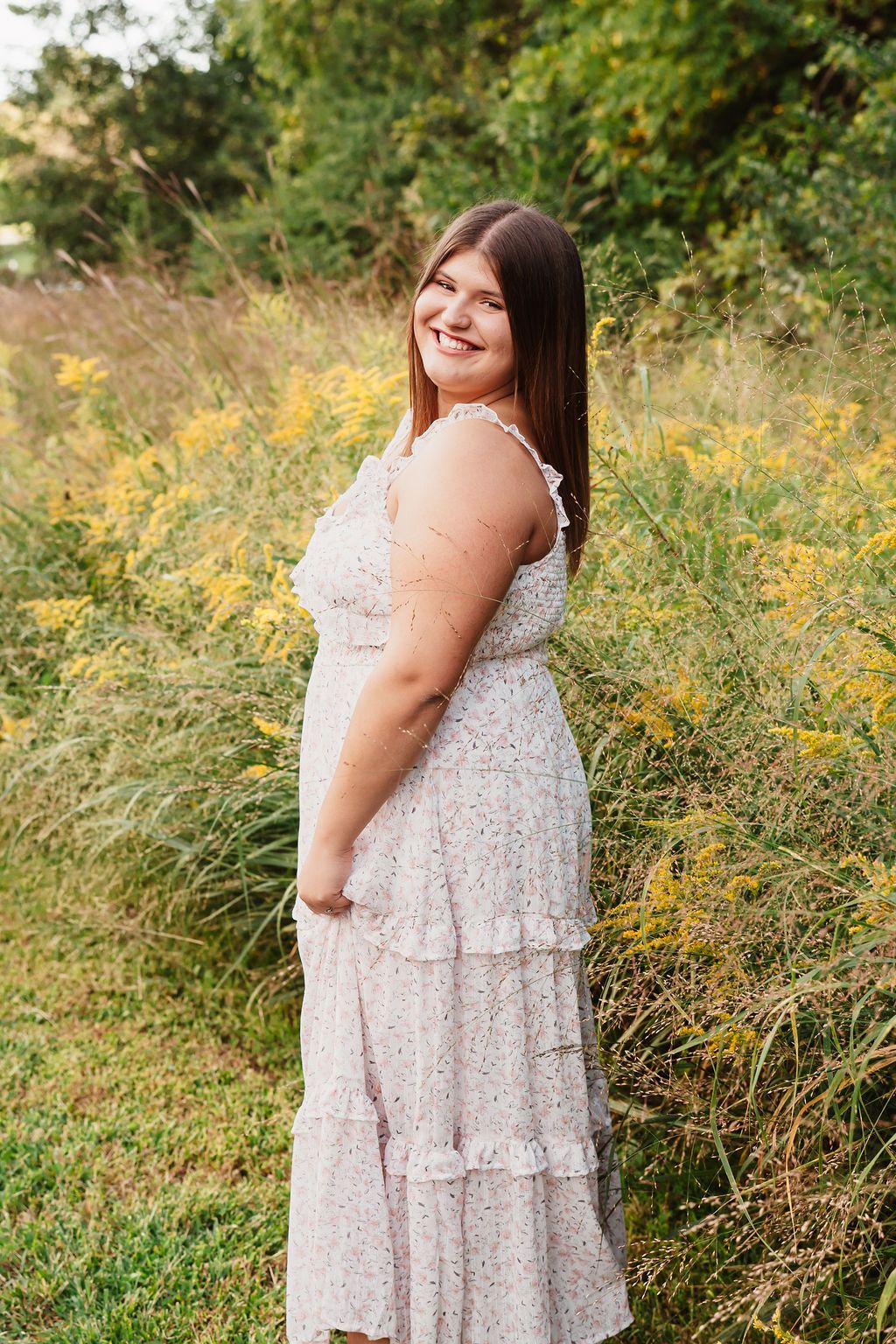 Smiling young woman in a floral tiered dress standing in a field of yellow wildflowers.