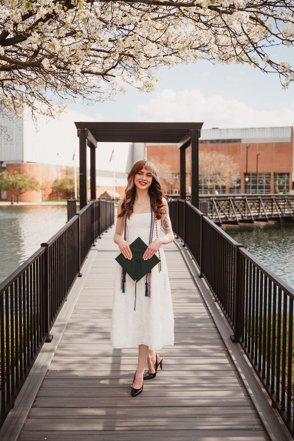 Smiling graduate in white dress holding green cap on wooden bridge with cherry blossoms and waterway in spring.