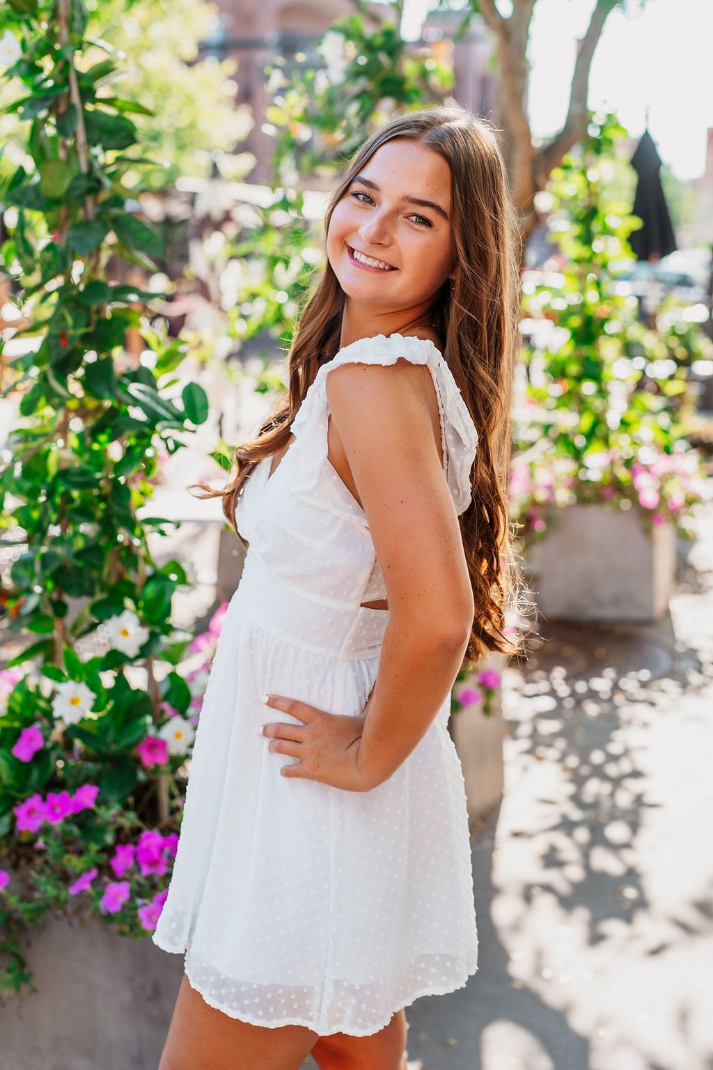 Smiling young woman in white dress posing outdoors among green plants and colorful flowers on a sunny day.