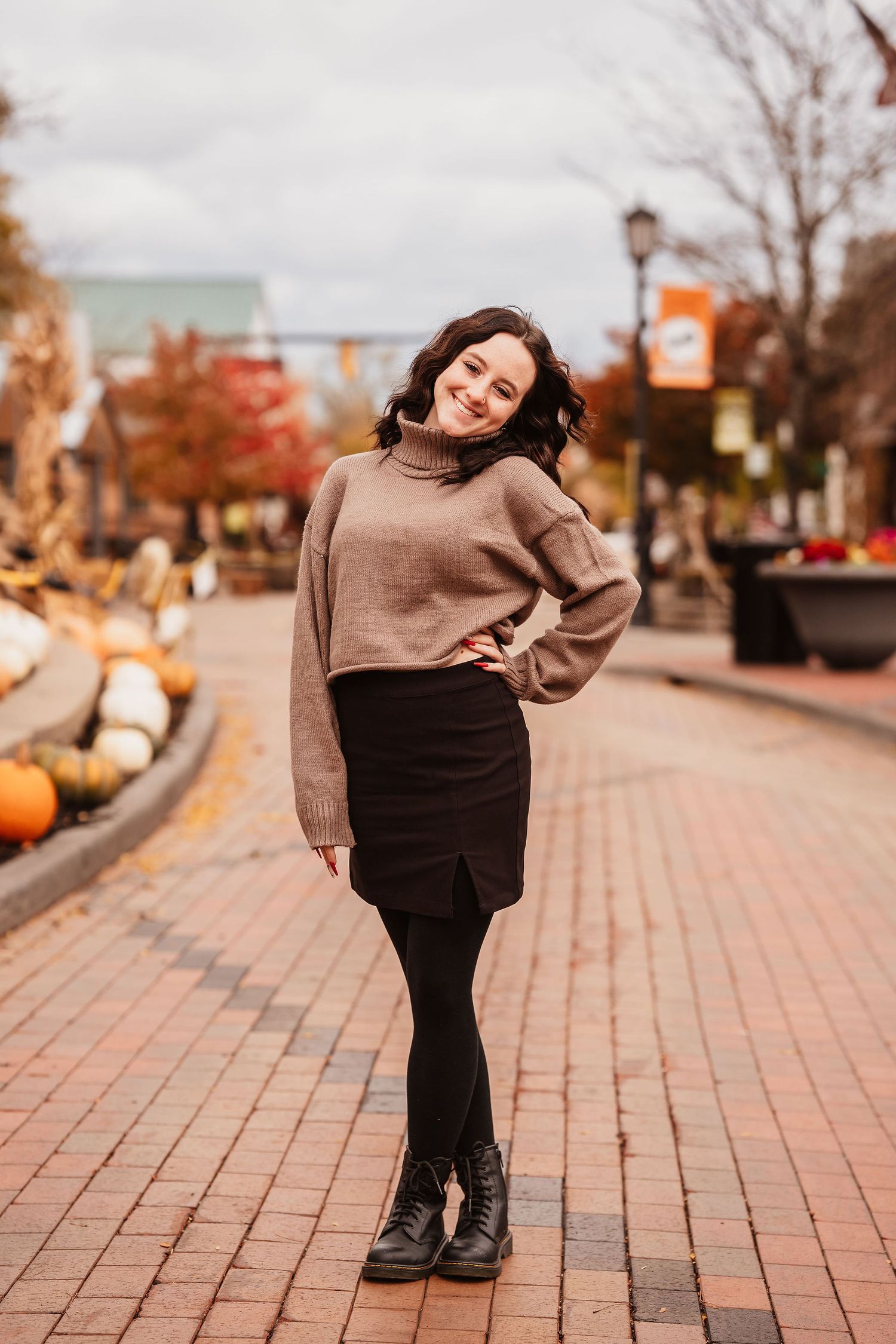 Young woman in tan sweater and black skirt smiles on brick path with fall foliage and pumpkins.