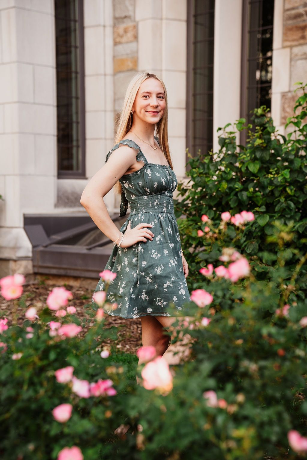 Blonde woman in green floral dress posing among pink roses outside a stone building.