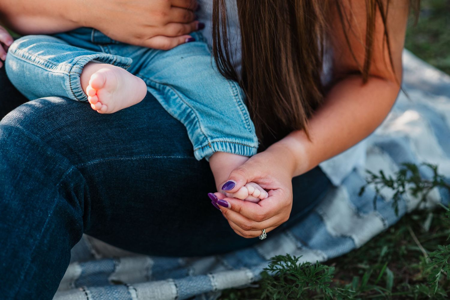 Mother sitting outdoors holding baby feet while painting tiny toenails purple on a blanket in the grass.