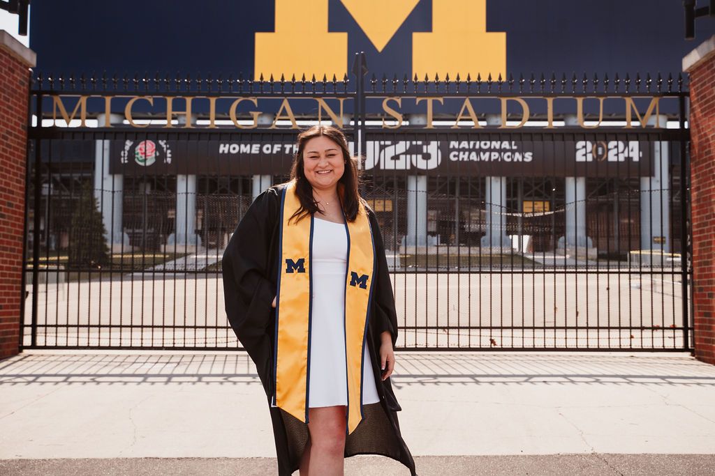 University of Michigan graduate in cap and gown posing in front of Michigan Stadium gates, celebrating 2024 National Champions.