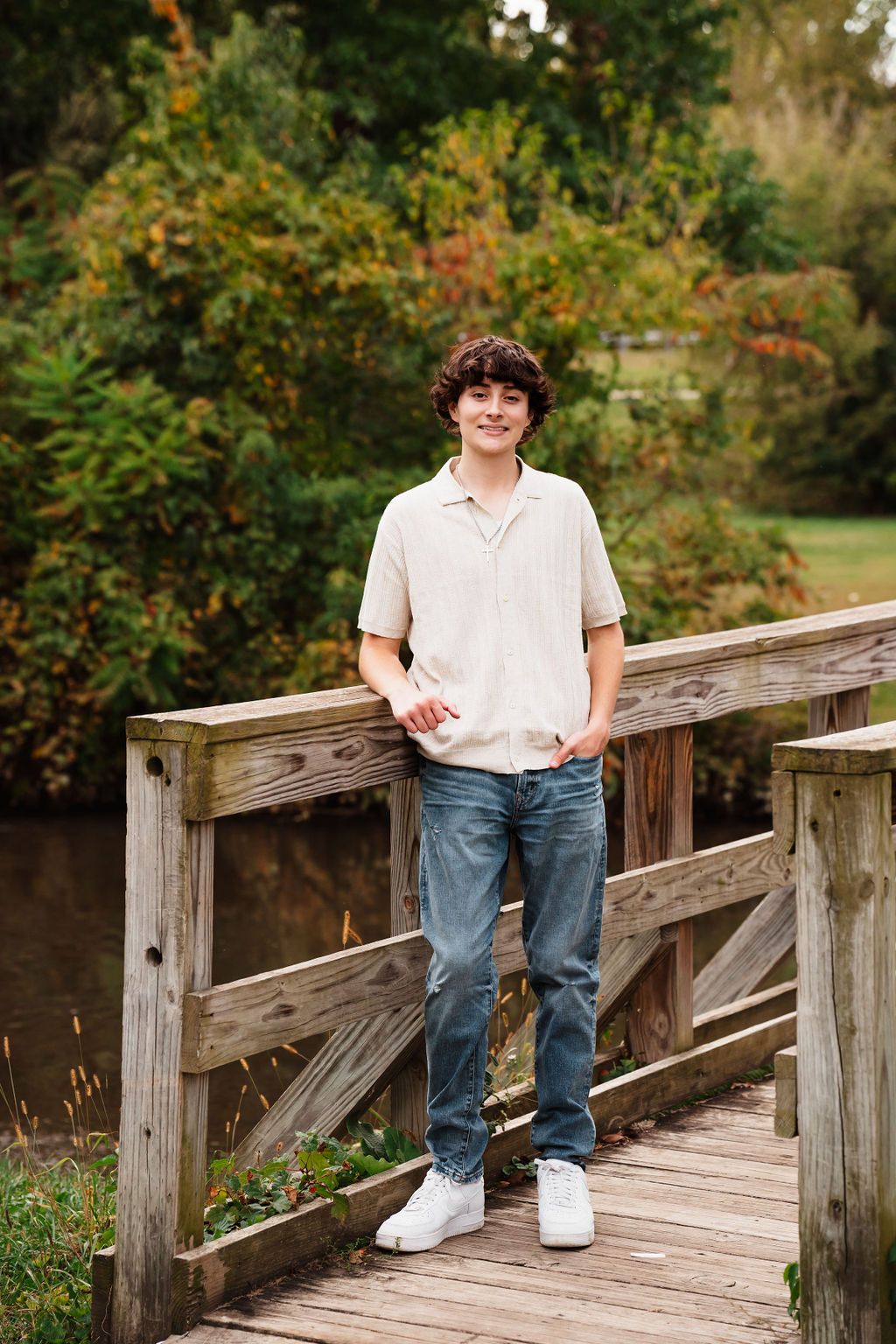 Smiling teen boy in beige shirt and jeans leans on wooden bridge railing in lush park setting.