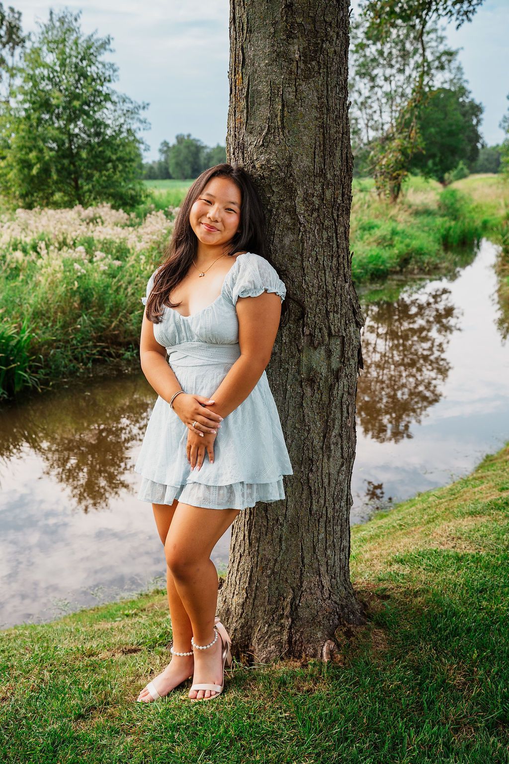 Person in light blue dress standing barefoot by tree near stream. Flower farm senior photos, Plymouth Michigan, PCEP Senior Photos