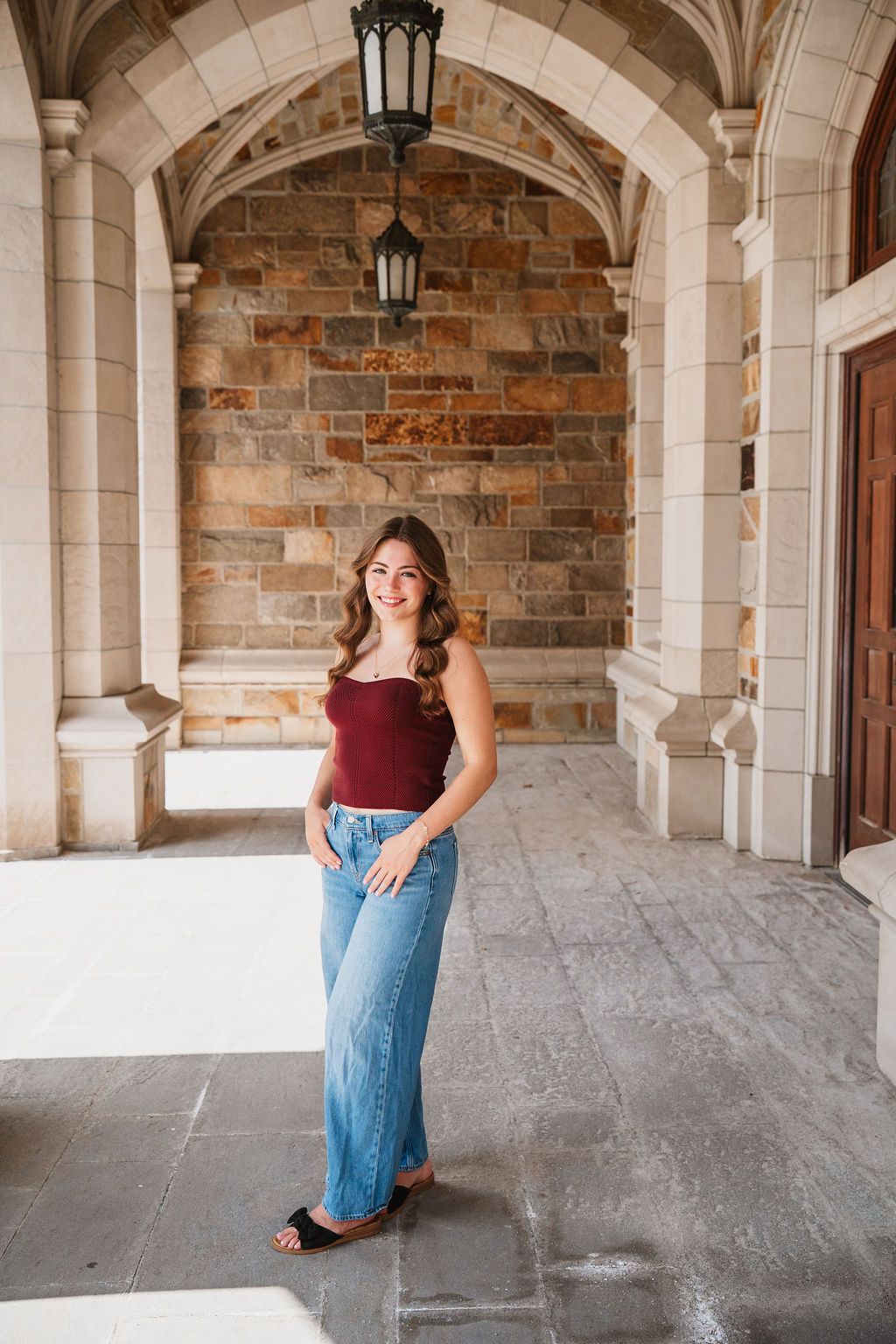 A person in a burgundy top and jeans poses in an elegant stone archway with natural lighting.