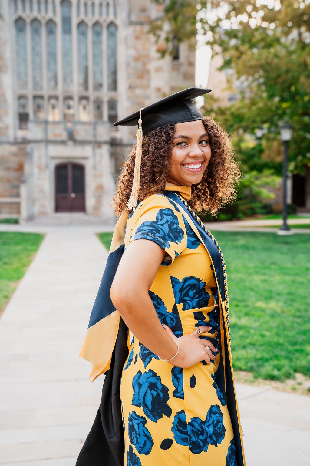 A graduate in cap and gown wearing a yellow and blue floral dress poses outside a Gothic university building.