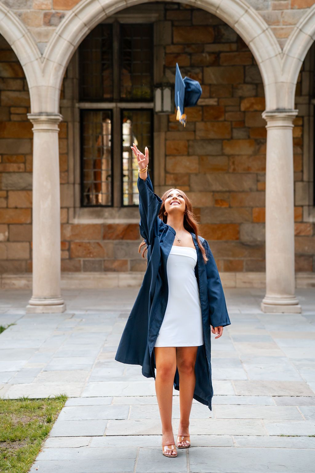 Graduate in blue cap and gown celebrates achievement in outdoor courtyard with stone arches and columns.