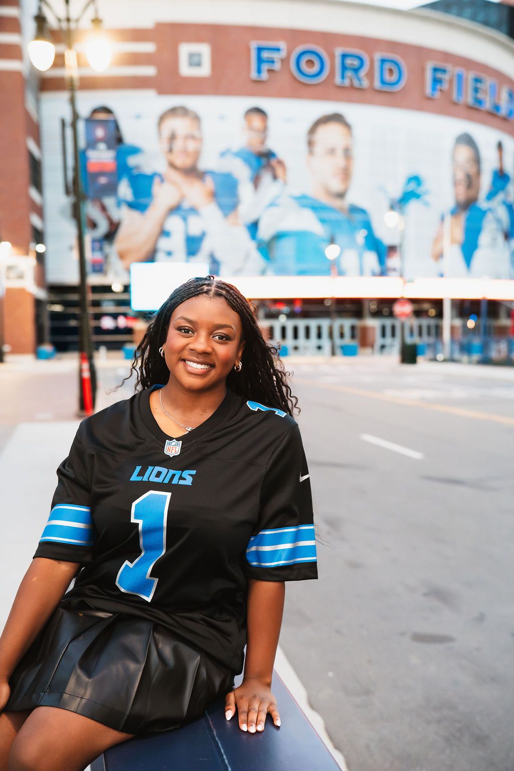 Detroit Lions fan poses outside Ford Field stadium wearing a black team jersey and smiling on game day.