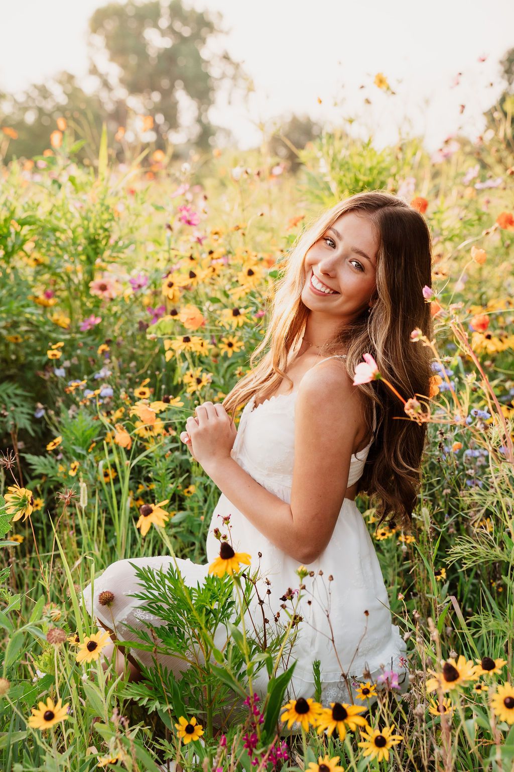 A person in a white dress sits among vibrant wildflowers during golden hour in a summer garden.