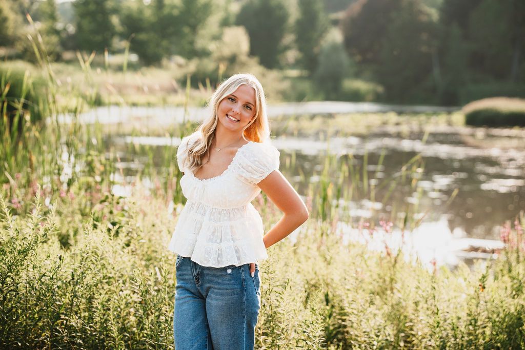 Young blonde woman smiling in white top and jeans, posing outdoors near a pond surrounded by lush green meadow plants.