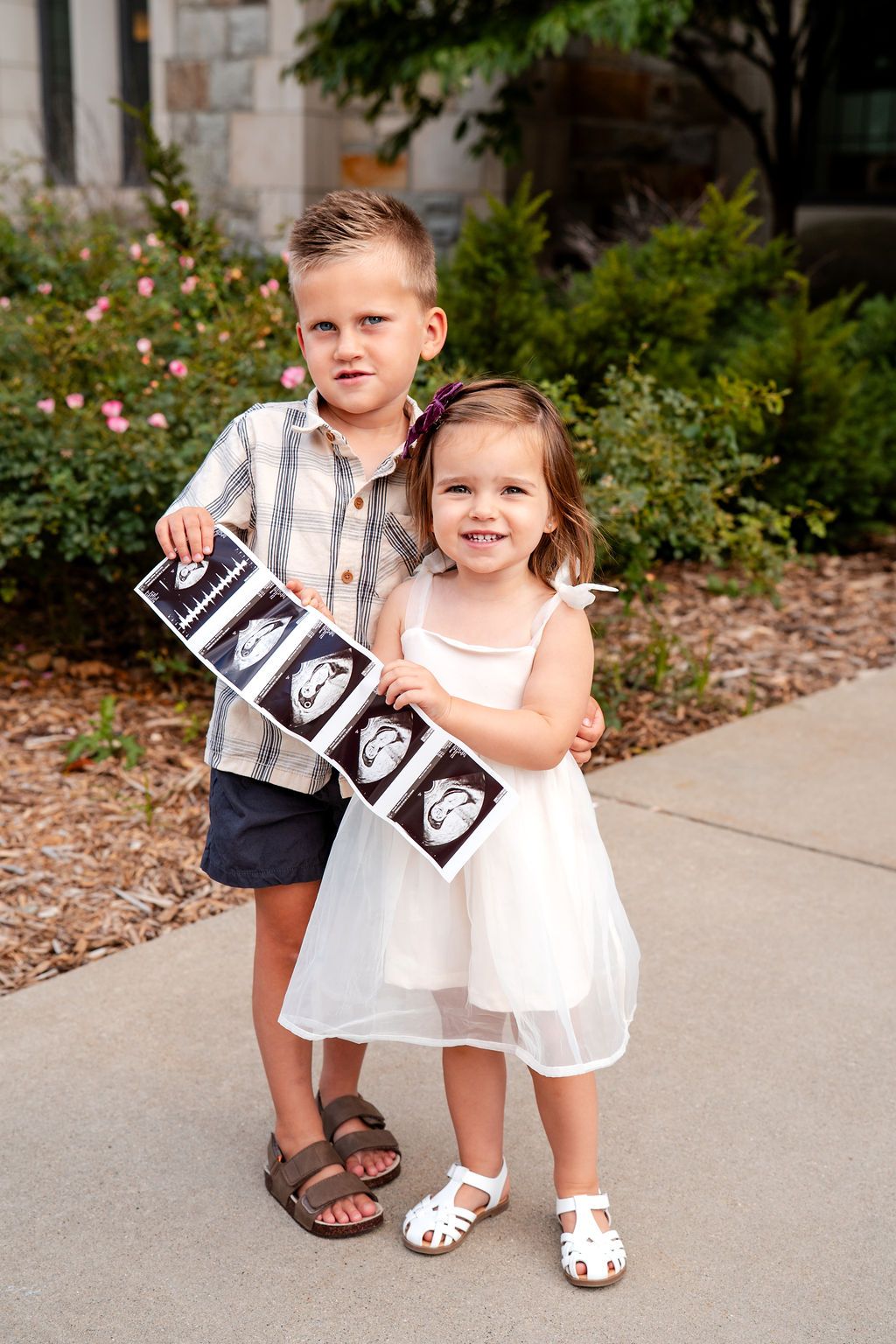 Two young children stand together holding ultrasound photos on a sidewalk near garden landscaping.