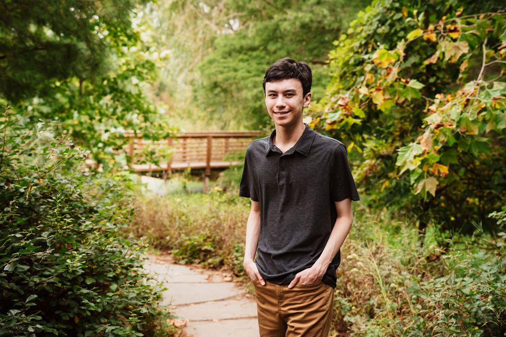 Young man smiling on a lush garden path with a wooden bridge and green foliage in the background.