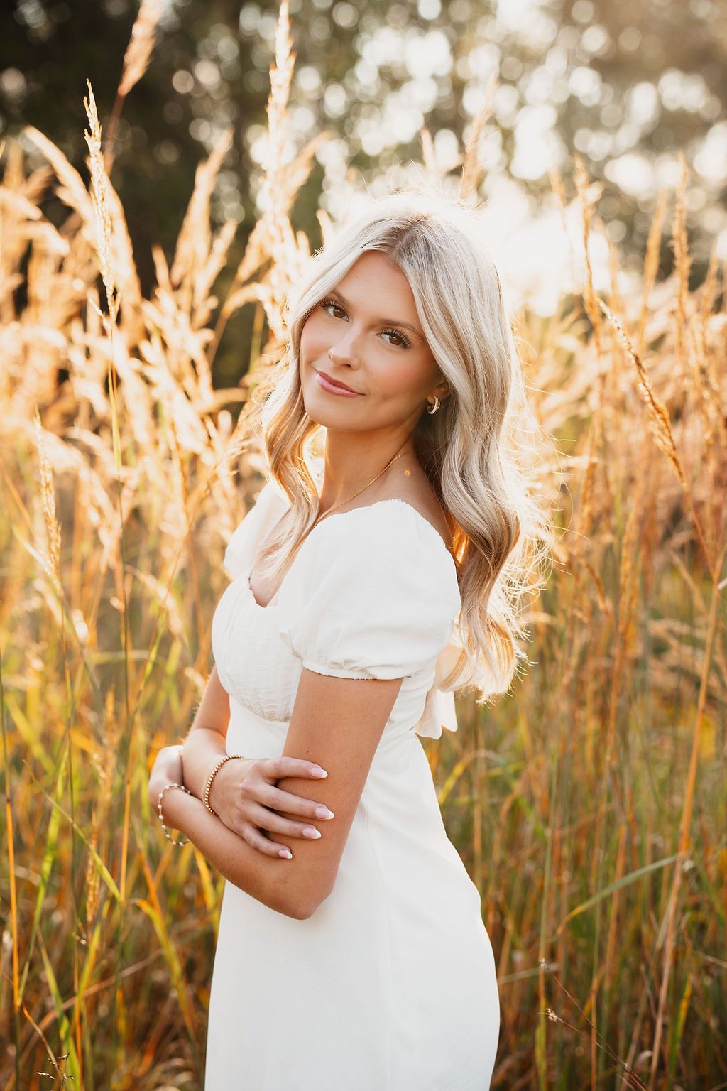A woman in a white dress stands in a golden wheat field at sunset with soft bokeh lighting effects in the background.