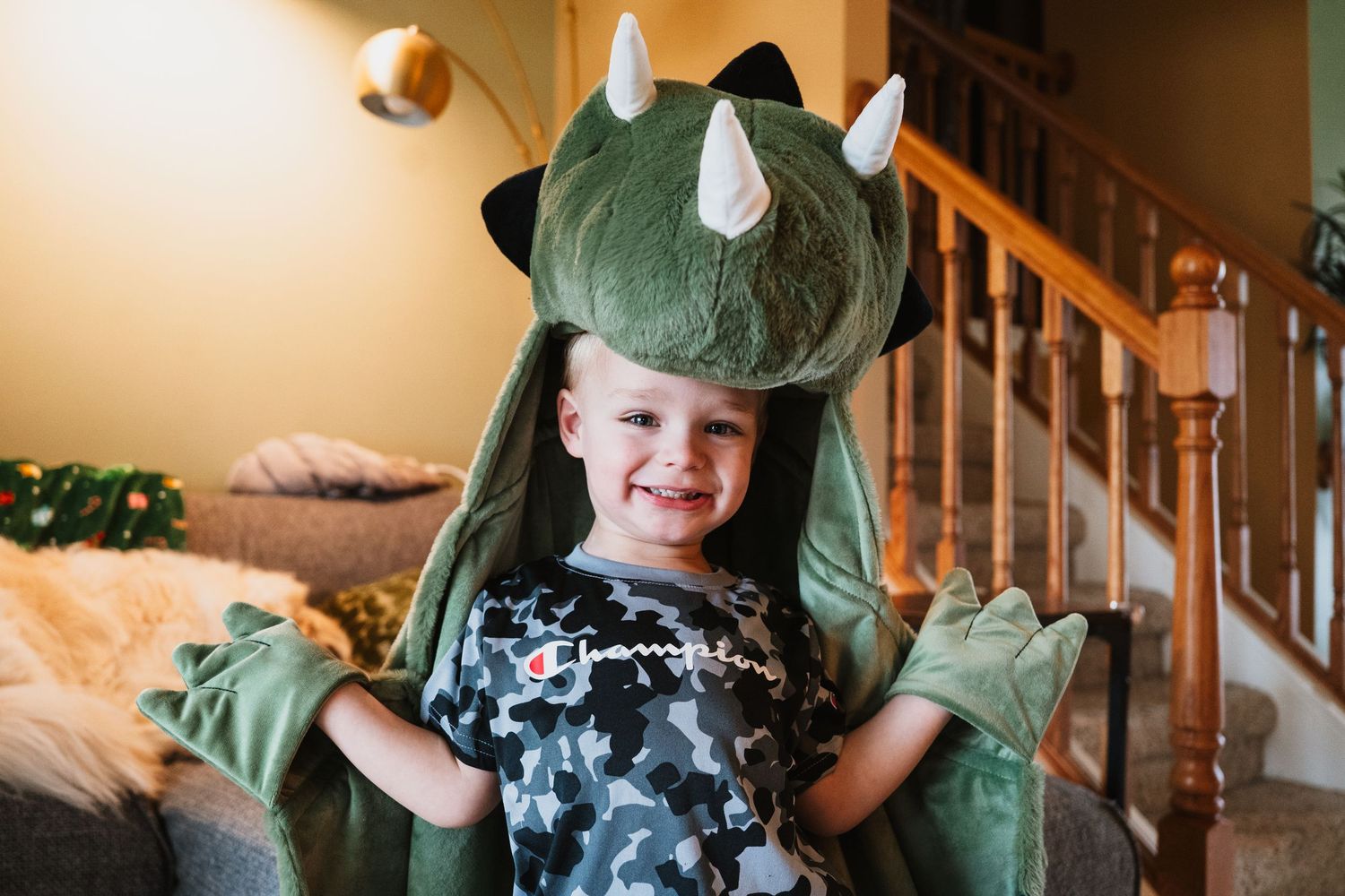Happy young boy wearing a green dinosaur costume with horns and wings, smiling indoors near a staircase.