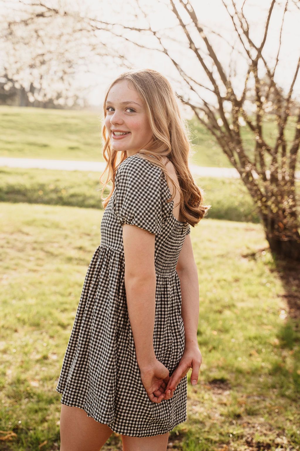 Young blonde woman in gingham dress posing outdoors near blooming tree on sunny day.