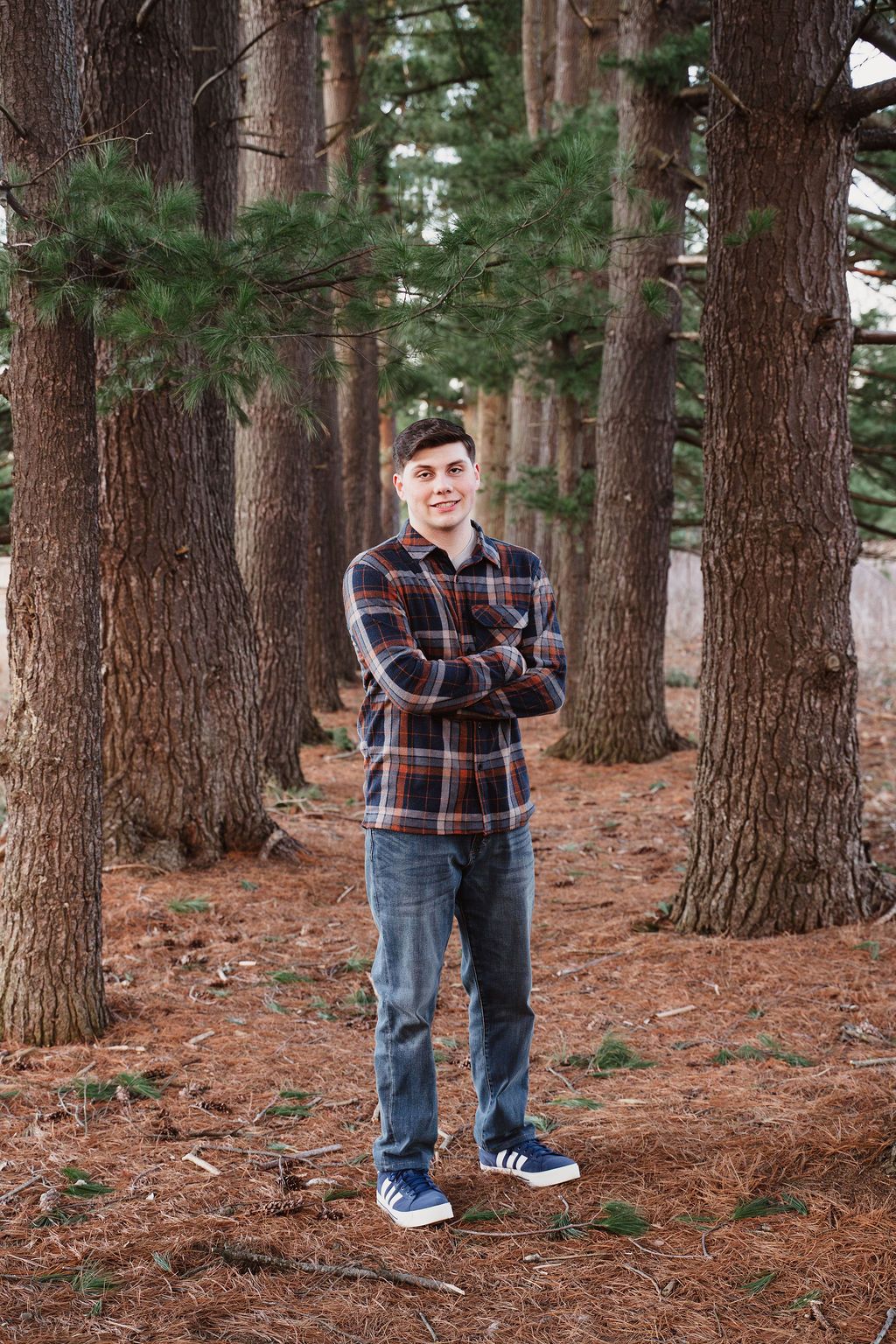 Young man in plaid shirt and jeans stands with arms crossed among tall pine trees on a forest floor covered with pine needles.