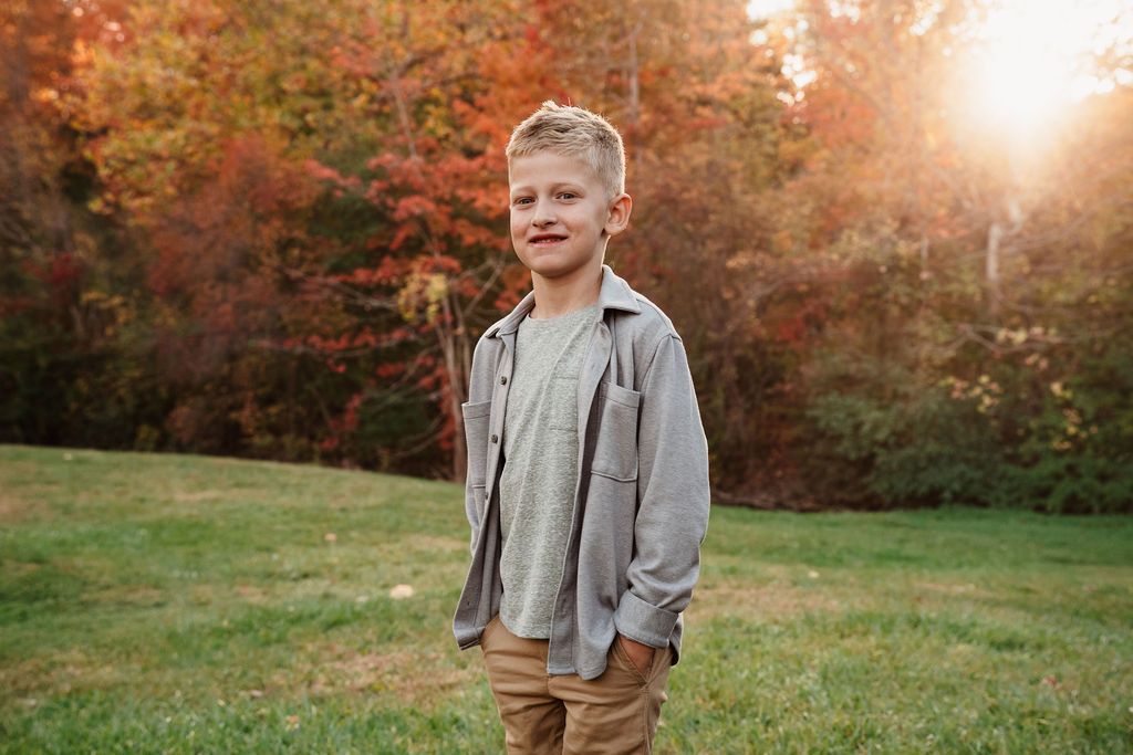 Smiling boy in gray shirt and khaki pants standing in a sunlit park with colorful autumn foliage.