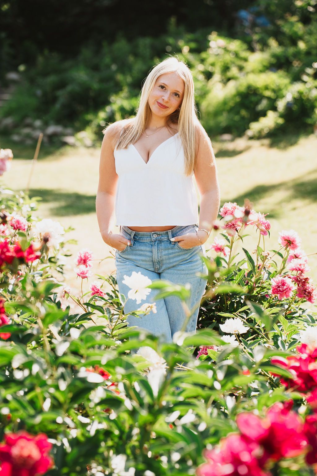 Blonde woman in white crop top and jeans posing among vibrant pink and white flowers in a garden.
