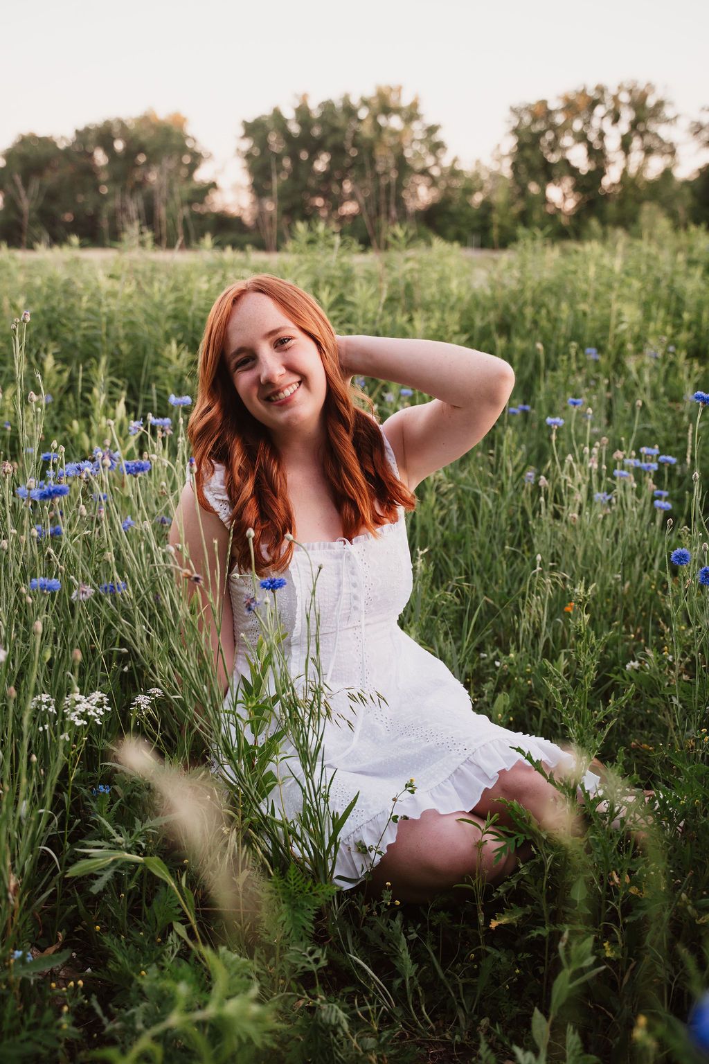 Young redhead woman in white dress sitting in wildflower field smiling, surrounded by blue cornflowers at sunset.