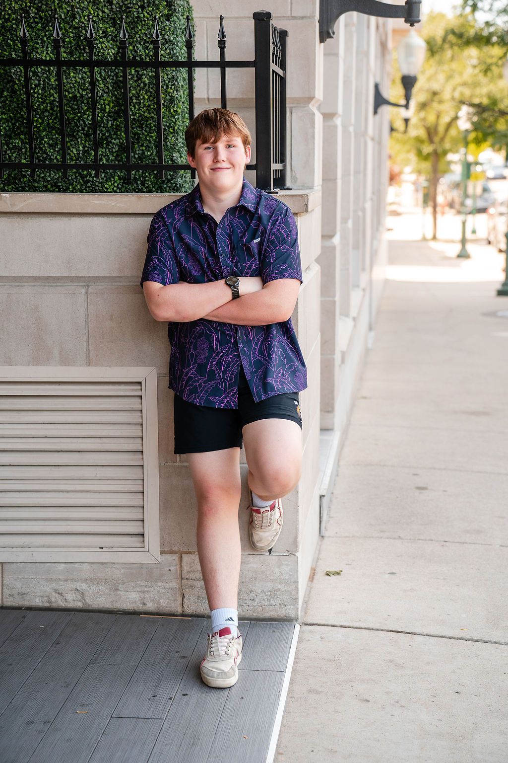 Teen boy in purple patterned shirt and black shorts poses confidently on urban sidewalk with arms crossed.