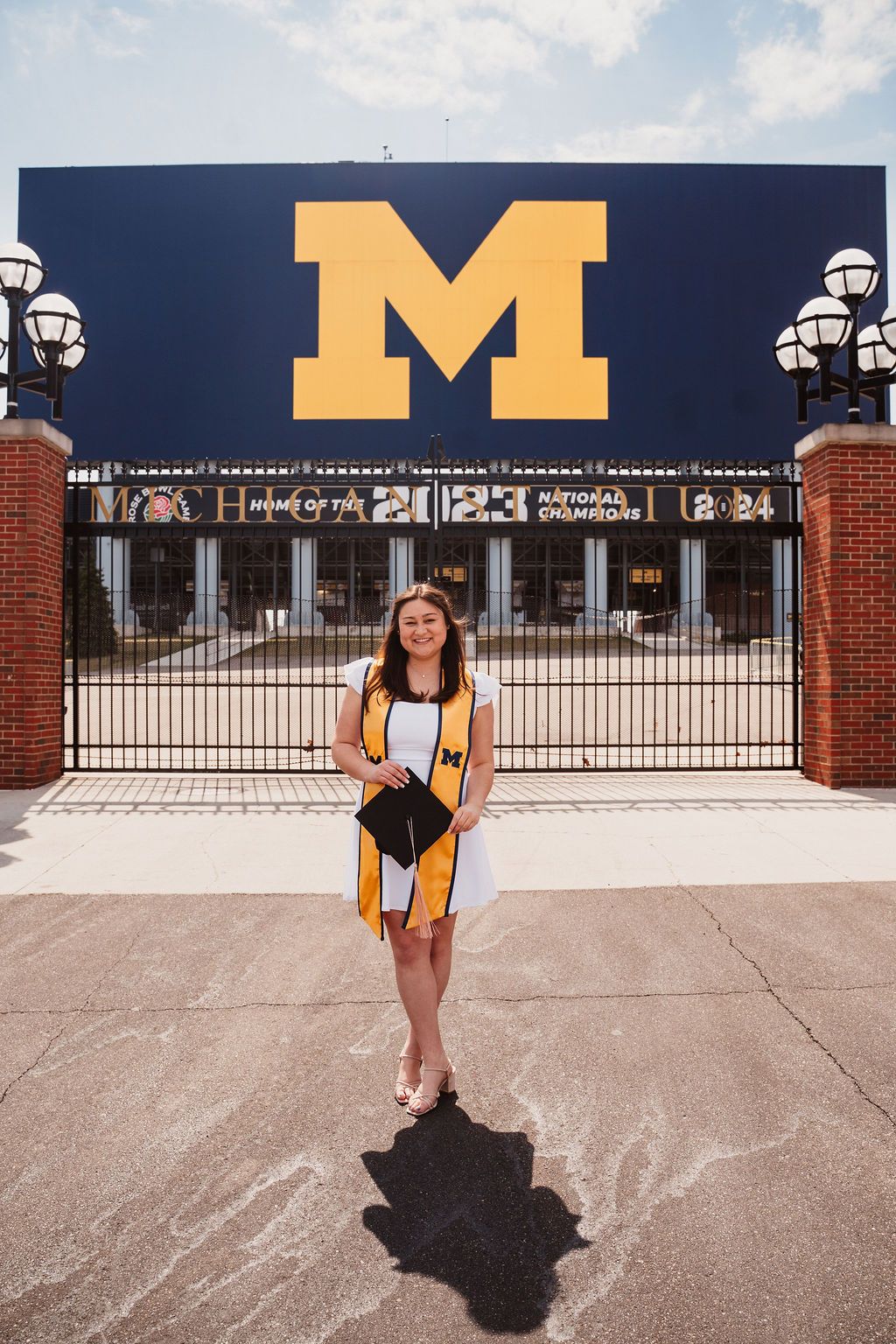 Smiling graduate in white dress with Michigan stole stands before iconic U of M stadium entrance.