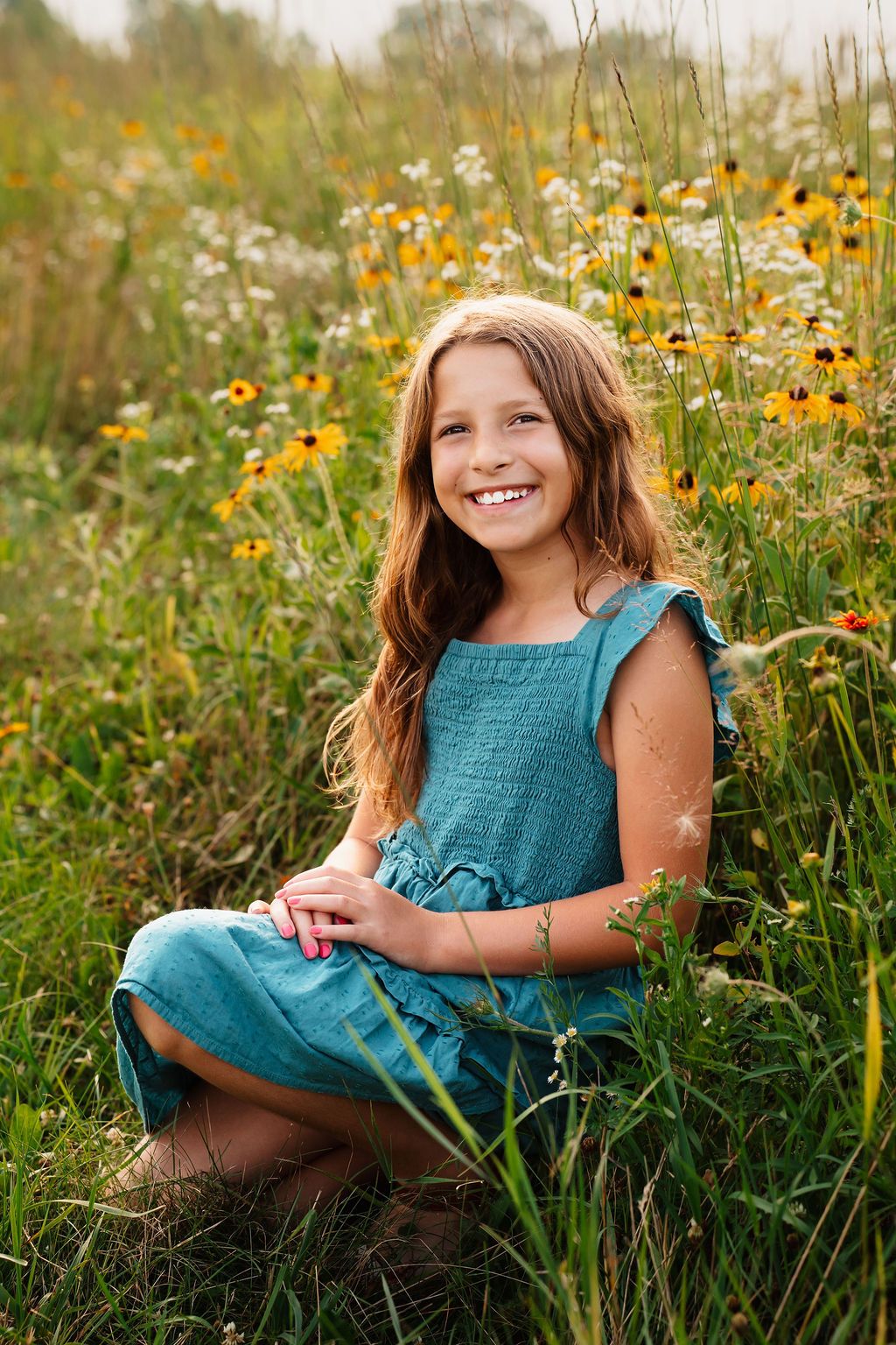 Smiling girl in teal dress sitting in wildflower meadow with yellow black-eyed susans and white flowers.