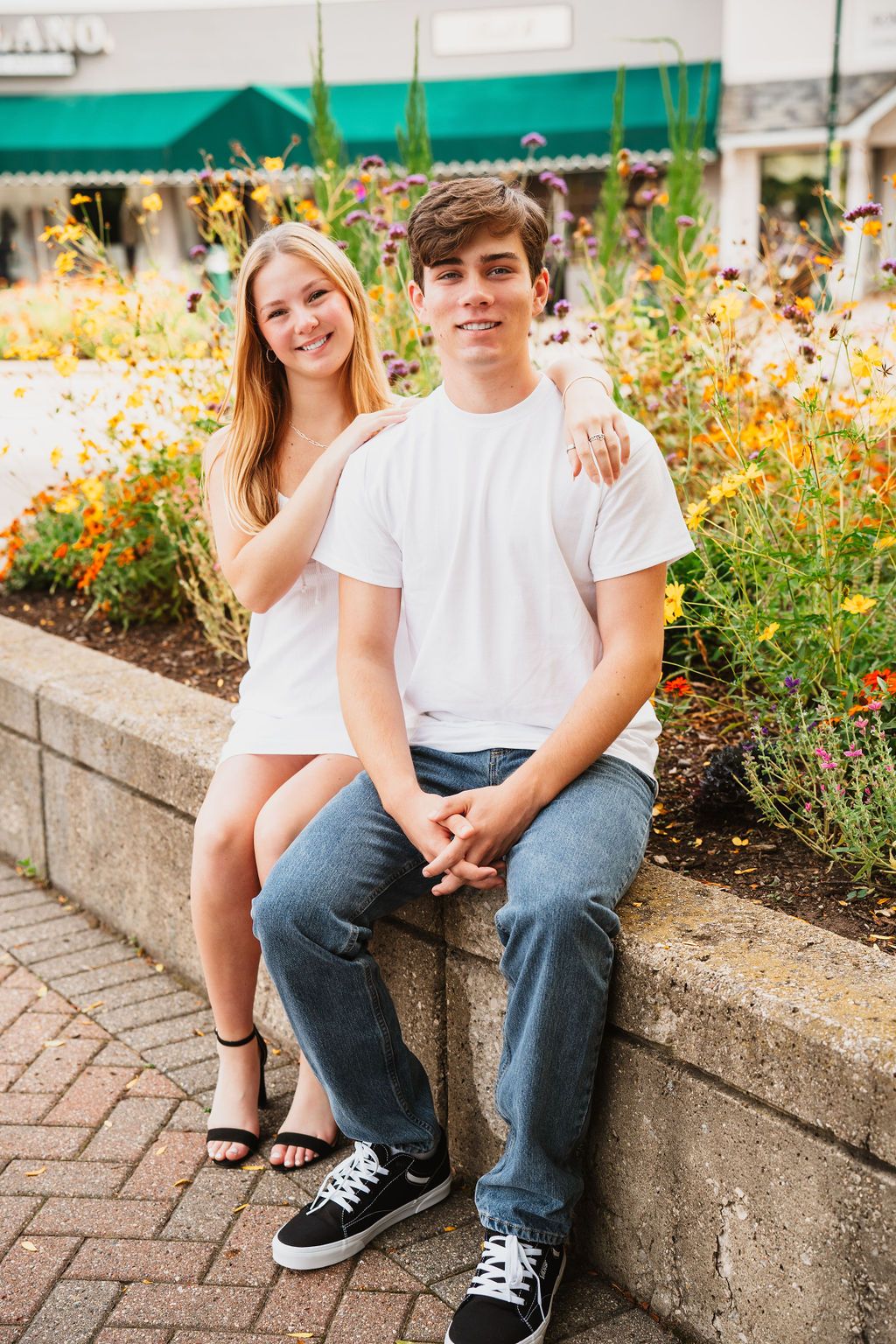 Teen boy and girl in matching white t-shirts and jeans sitting together on a stone wall with colorful flowers behind them.