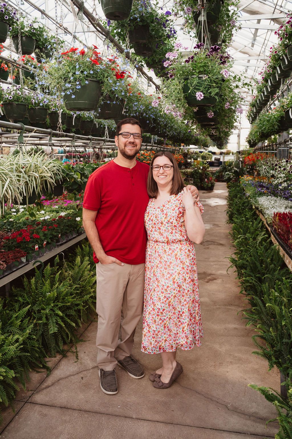 Couple smiling together in a lush greenhouse surrounded by colorful hanging flowers and green plants.