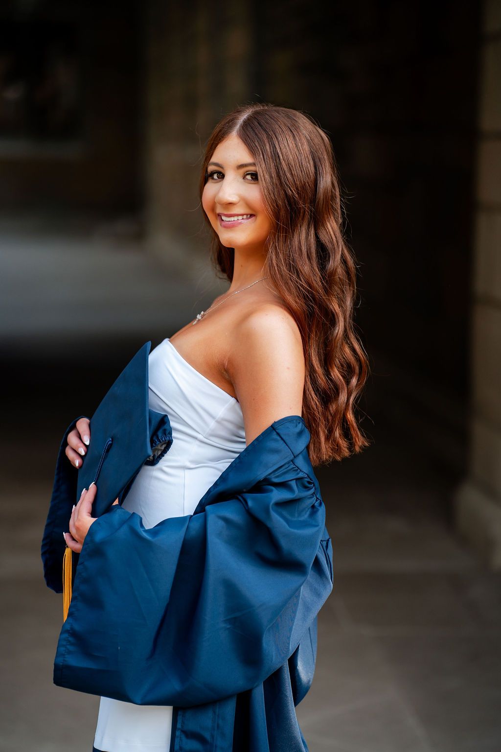 Graduate in white dress and navy blue graduation gown poses outdoors while looking over shoulder with flowing brown hair.