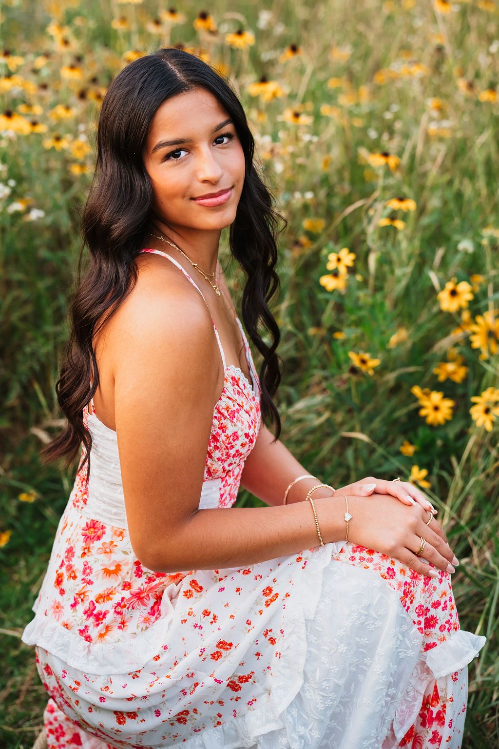 A person in a white and red floral dress poses gracefully in a field of yellow wildflowers on a sunny day.